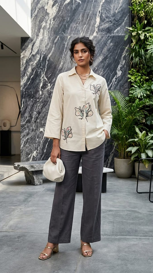 Woman wearing a beige embroidered shirt and gray pants in an indoor setting with plants and marble wall.