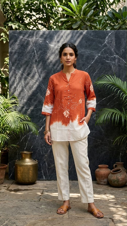 Woman wearing an orange and white patterned top with white pants standing in front of a dark wall with plants.
