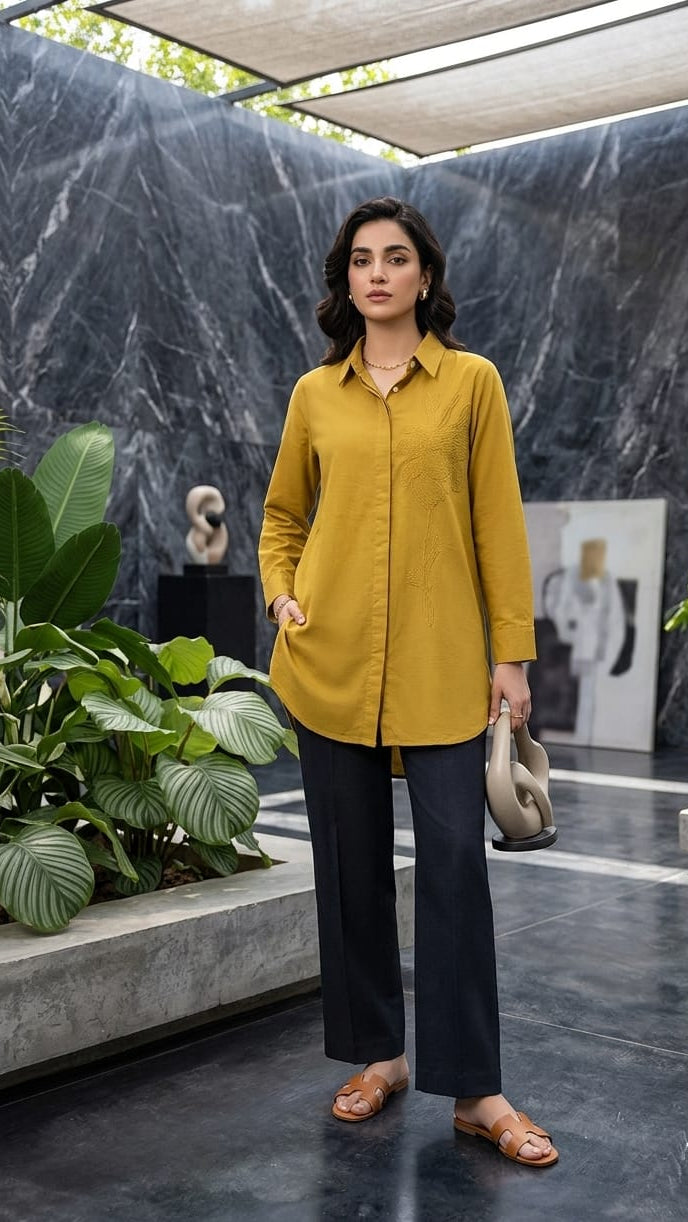 Woman in a mustard yellow shirt and black pants standing in a modern indoor setting with plants and abstract art.